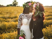 A Black human in a wedding dress and white human in a suit celebrate their wedding day, leaning in for a kiss.