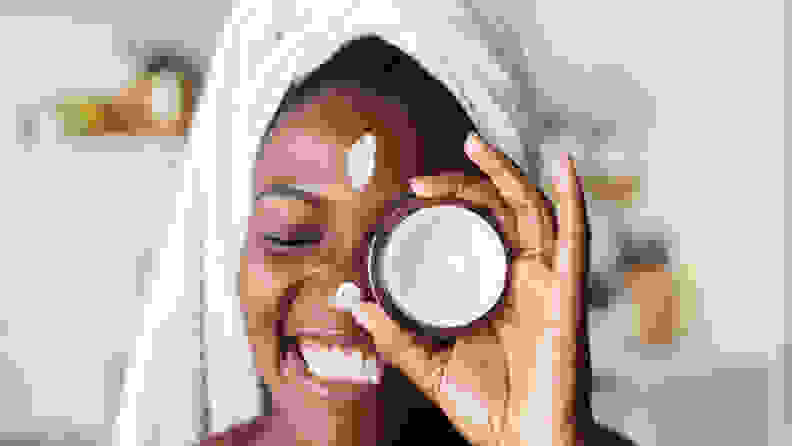 A person holds up a jar of moisturizer to their eye and smiles.