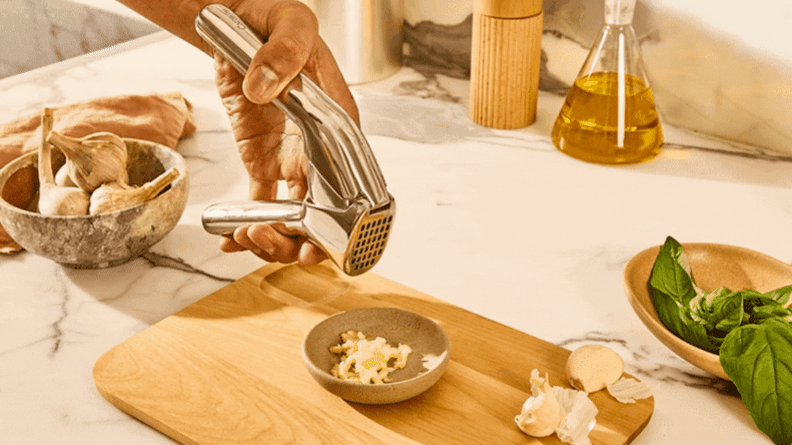 Person squeezing fresh garlic out of garlic press into small dish on top of wooden cutting board.