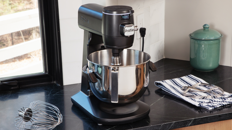 The GE Profile Smart Mixer sits on a black counter in front of a window with the whisk attachment to its left and the paddle attachment on a dish towel to its right.