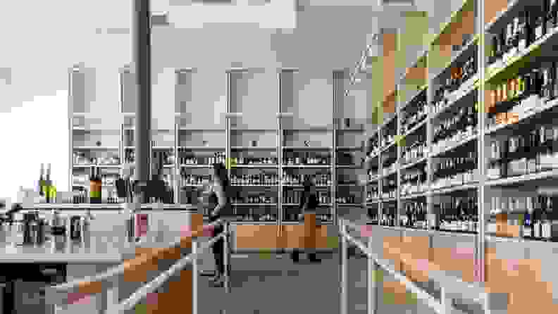 Four people shop for wine in a brightly lit natural wine store with light wood shelves filled with colorful bottles.