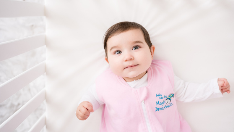 A baby wearing a pink onesie lays in a crib.