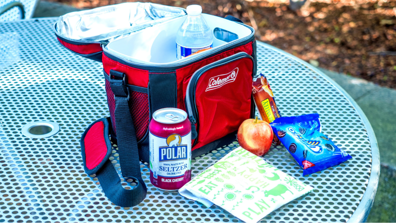 On an outdoor picnic table, there's a half-opened Coleman lunch cooler, with a can of soda, a lunchskins sandwich bag, an apple, and Oreos next to it.