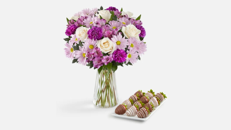 A bouquet of white roses, purple carnations, and pink daisies in a glass vase next to a plate of chocolate-covered strawberries.
