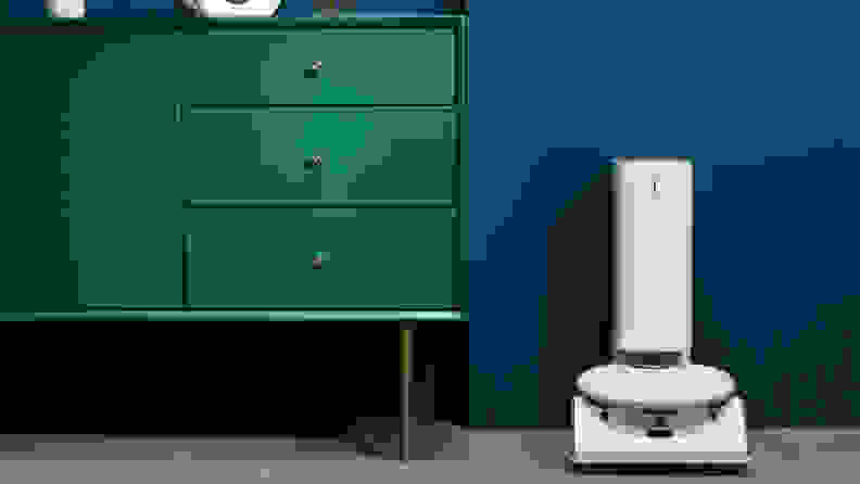 A white robot vacuum sits in its doc next to a green dresser