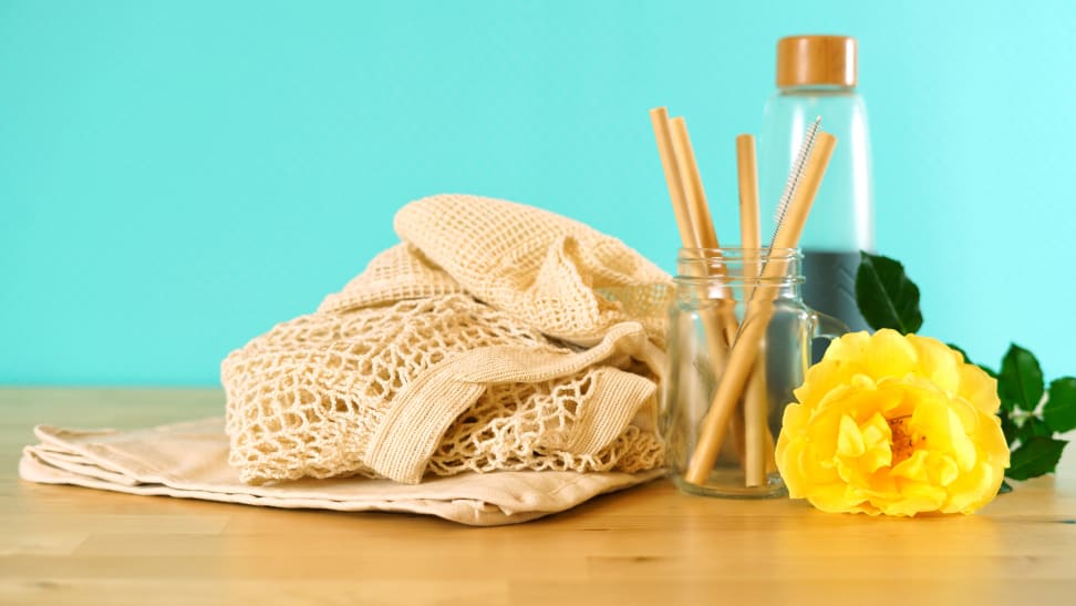 A reusable bag, straws, and glass container and yellow flower against teal background.