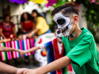Small children with skull painted faces playing together while holding hands in a circle at a Dia de Los Muertos event outdoors.