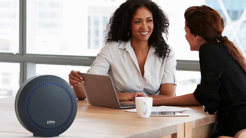 An air purifier sits on a table while two people work.