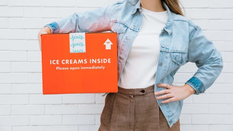 A person holds a bright orange cardboard box with the Jeni's Splendid Ice Creams logo on it.