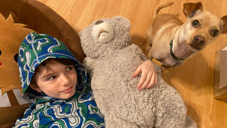 A child holds his Squishmallow and teddy bear a he looks at his puppy.