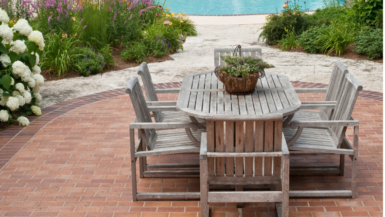Wood patio set with plant sitting on brick patio flooring, surrounded by shrubs and flowers
