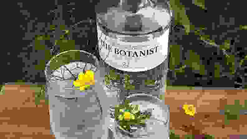 A bottle of gin sits on a wooden tables outside accompanied by two mixed drinks, garnished with yellow flowers.