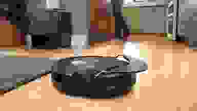 A black, round robot vacuum on a light wooden floor next to a gray rug, with a person standing in the background