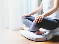 A person in yoga pants sits cross-legged on a chair cushion, which for the photo is positioned on a hardwood floor.