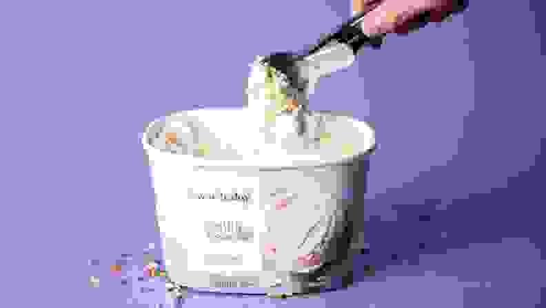 A hand scooping into a tub of vanilla ice cream on a purple background, surrounded by rainbow sprinkles.