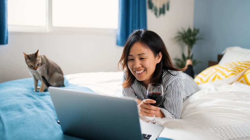 A person lying in bed made with white sheets and holding a glass of red wine while watching a movie on a laptop