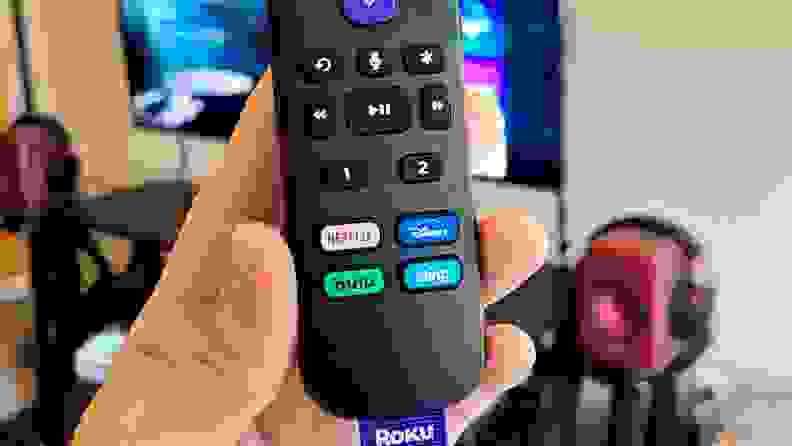 A hand holds the black, rounded remote in front of a TV console showing keys, including programmable ones as well as quick keys for streaming services.