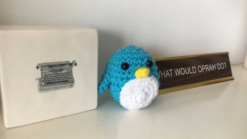 A blue crocheted penguin toy sits on a desk next to a typewriter paperweight and a sign that says 