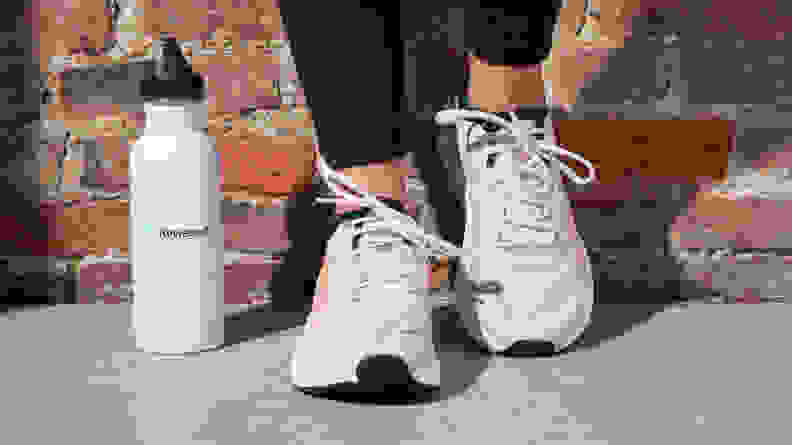 A person leans against a brick wall wearing the Puma Run XX Nitro running shoes standing next to a white water bottle.