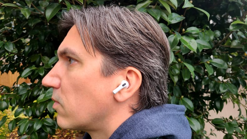 The all-white AirPod sits in the reviewer's ear with its shortened stem facing forward, set above a gret sweatshirt hood and below brown grey hair.