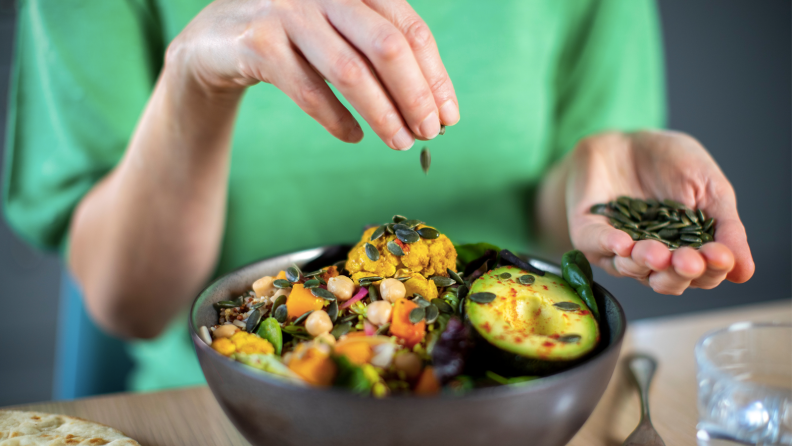 A person sprinkles seeds into a bowl of salad from just out of frame.