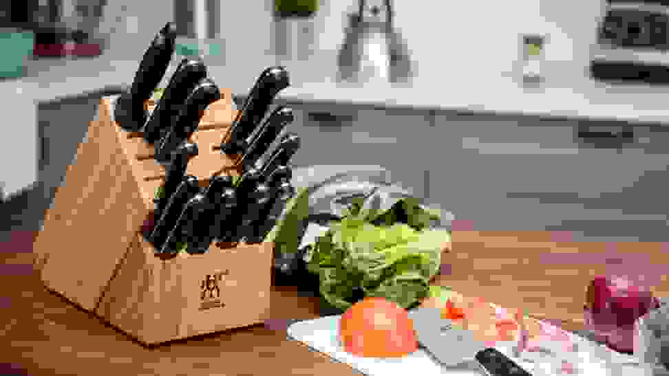 A wooden knife block filled with assorted kitchen knives on a wood counter next to a plastic cutting board and chopped produce.