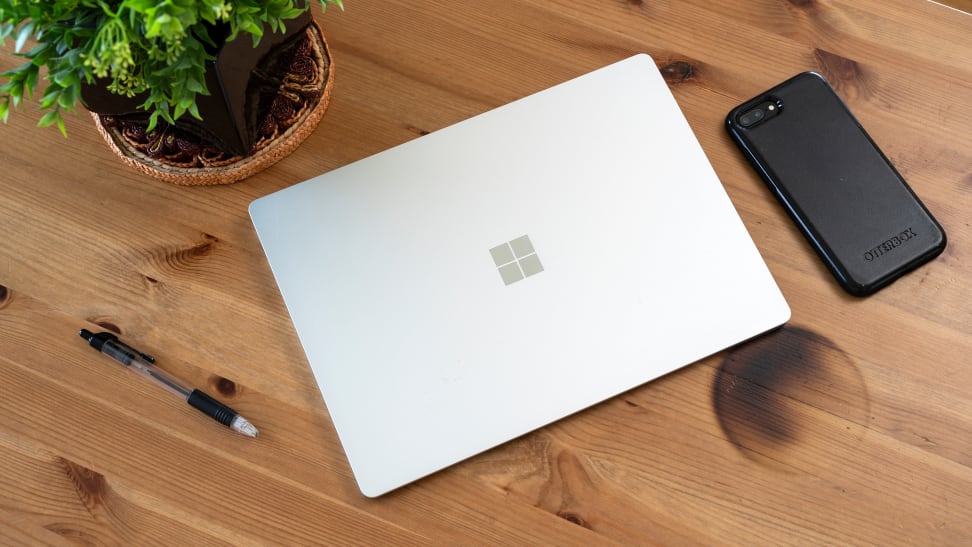 A Microsoft Surface 3 laptop on a brown table. Besides the laptop are a smartphone, a pen, and an indoor plant.