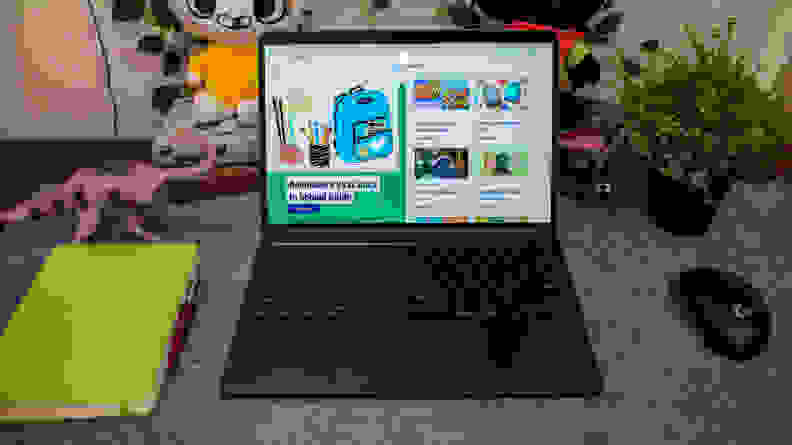 An open and powered on laptop on a grey desk mat with a green note book to the left and a black mouse to the right