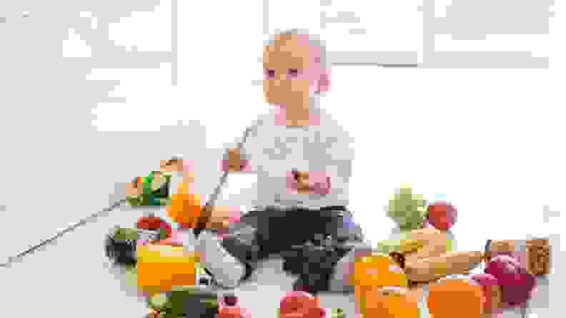 A baby sits on the floor surrounded by whole fruits and vegetables, as it clutches a wooden spoon.