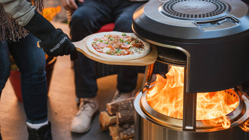 Person wearing gloves uses wooden pizza peel to insert uncooked pizza inside of Solo Stove Pi Fire pizza oven with open flame.