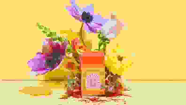 An artistic lifestyle image featuring a box of saffron and various purple, green, and yellow flowers sprouting out from behind it.