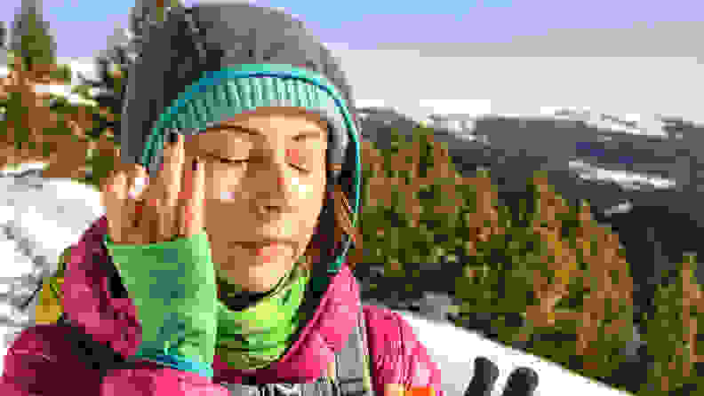 A person in winter gear outside applying sunscreen to their face.