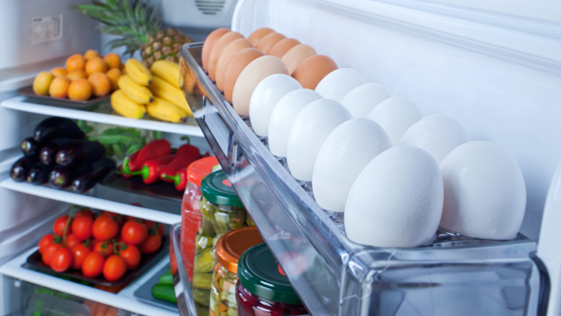 The inside of an open fridge that's fully stocked with eggs, fruits, and vegetables