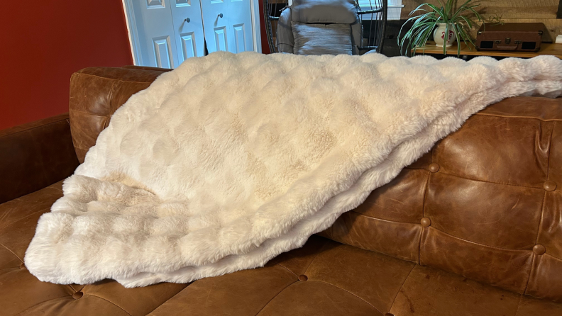 A cream colored Cozy Earth blanket is tossed across a brown leather couch.