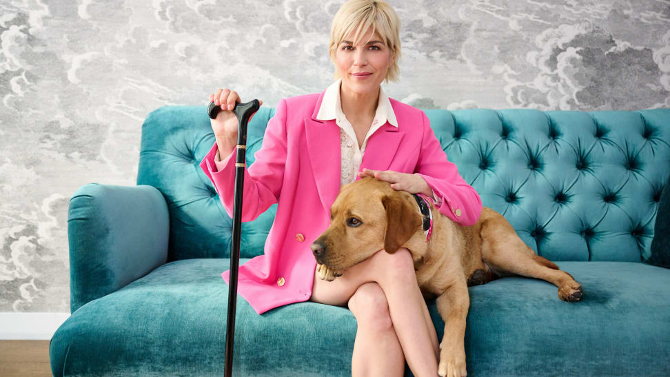 Selma Blair, the QVC Brand Ambassador for Accessibility, sits on a teal velvet sofa wearing a pink blazer and white button-down while holding a cane with a dog resting on her lap.