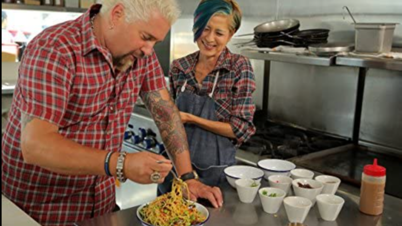 A still of Guy Fieri trying food in a kitchen.