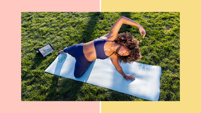 A woman doing a pilates stretch on a blue Alo Move yoga matt outside, on the grass.
