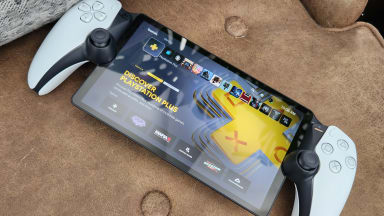 A handheld gaming console, the playstation portal, on top of a brown chair table with a gray pillow next to it