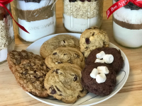 A plate of four kinds of cookies sits on a wood counter in front of jars of cookie mix tied with red ribbons.