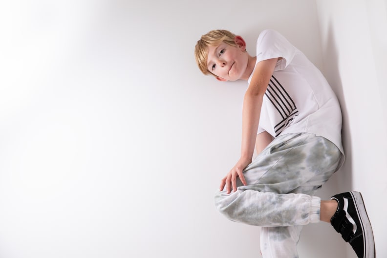 A boy wearing a white T-shirt, light green tie dye pants and black tennis shoes leans against a wall.
