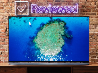 An OLED TV with an island on screen sits on a wooden desk before a brick wall with the neon sign that says "reviewed."