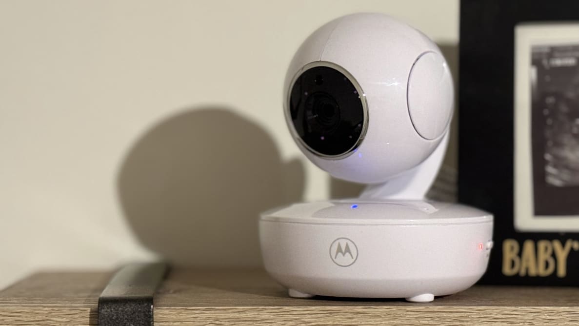 A Motorola VM36XL baby monitor camera sits on a wooden shelf next to a picture frame.