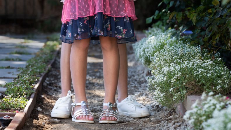 A pair of children's feet wearing silver sandals in front of a pair of children's feet wearing white tennis shoes.
