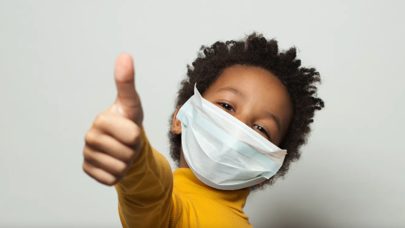 Child wearing face mask happily holding a thumbs up sign.