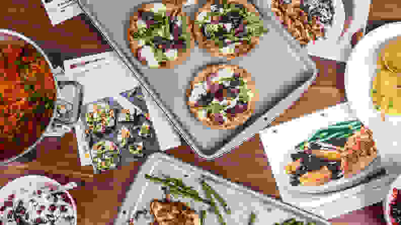 Various dishes are being prepared on a table alongside pages from a recipe book. At the center of the photo is a pan of small homemade pizzas.