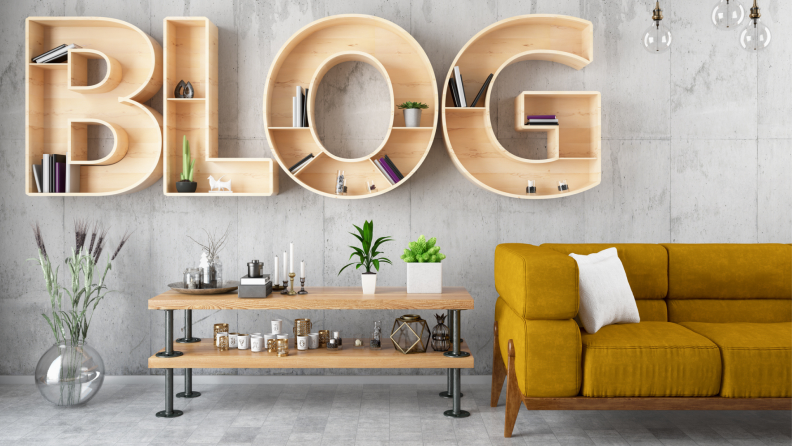 An office interior has been professionally decorated with plants, candles, and other knickknacks arranged on a wooden table with two tiers. To the right of the table, there is a gold-colored couch. Shelving units hung on the wall are shaped like letters, spelling out the word “blog.”