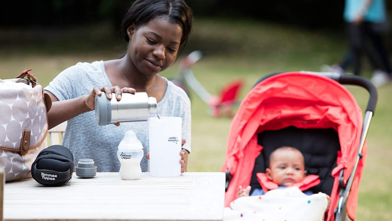A mother using a portable bottle warmer on a picnic table with her baby next to her in a stroller.