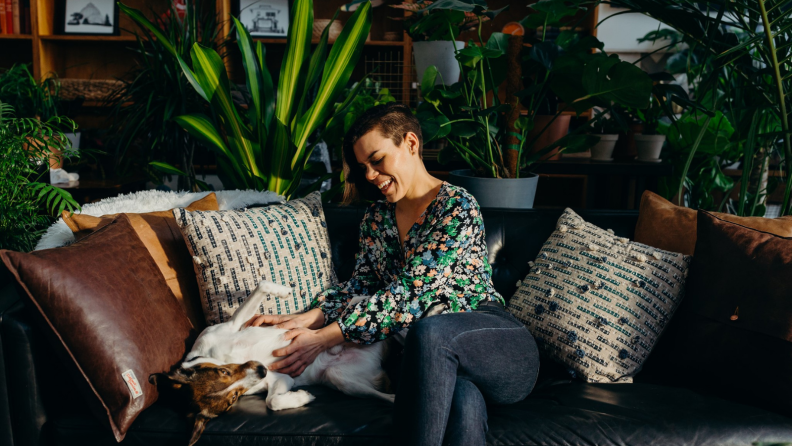 A woman pets her dog in a room of plants.