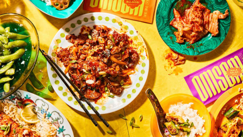 On a yellow table, Southeast Asian dishes are on display with the Omsom sauce kits around.