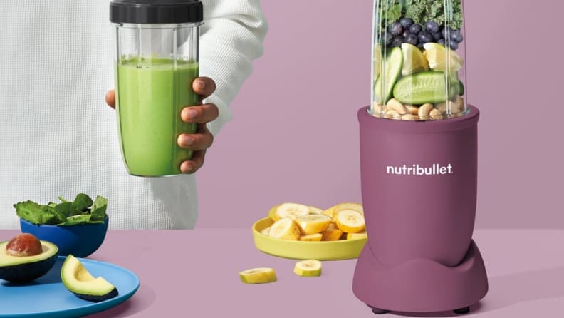 A pastel purple personal blender filled with fruit next to a hand holding a blended green smoothie.
